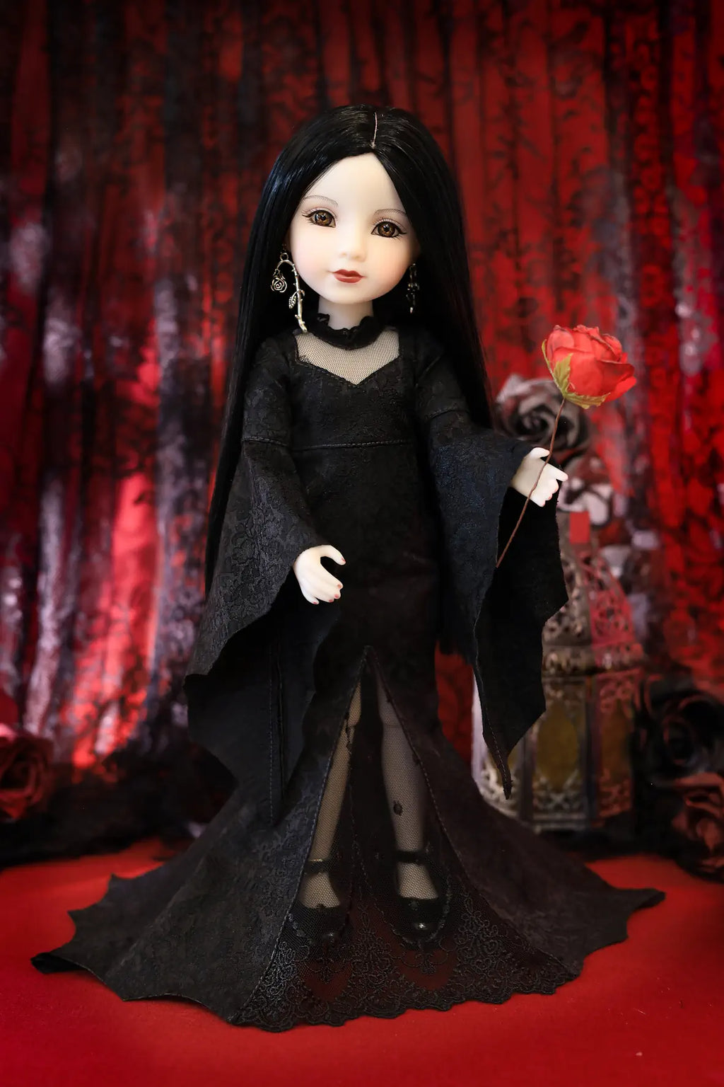 A gothic-style doll with long black hair, pale skin, and a black lace dress, holding a red rose against a dark red patterned curtain backdrop.