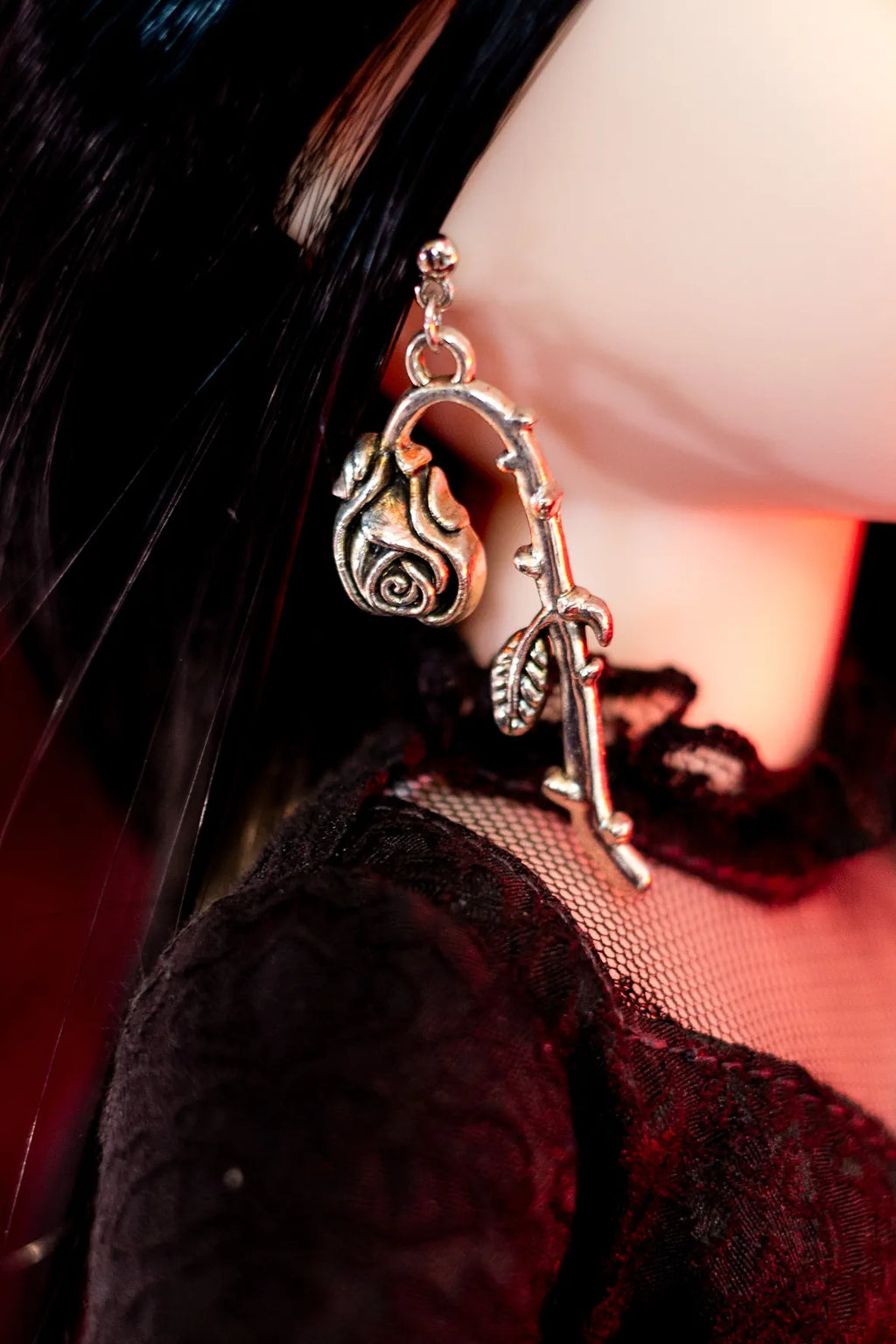 A close-up of a doll wearing a dangling earring shaped like a rose on a branch, with black hair and a black lace garment visible against a dark background.