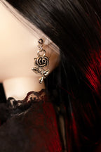 A close-up of a gothic-style rose earring with a silver-toned metal finish, shown on a doll with long black hair and a black and red lace top.
