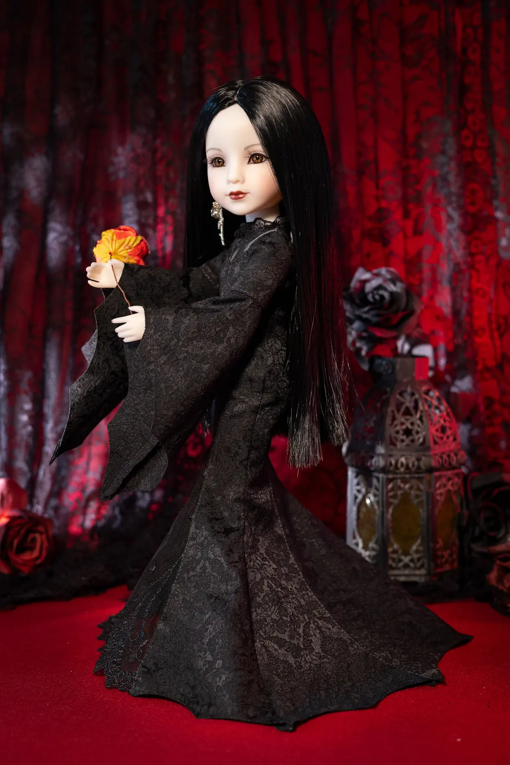 A doll with long black hair and pale skin wearing a long black dress, holding a red and orange flower, posed against a dark red patterned curtain backdrop with a lantern and roses.