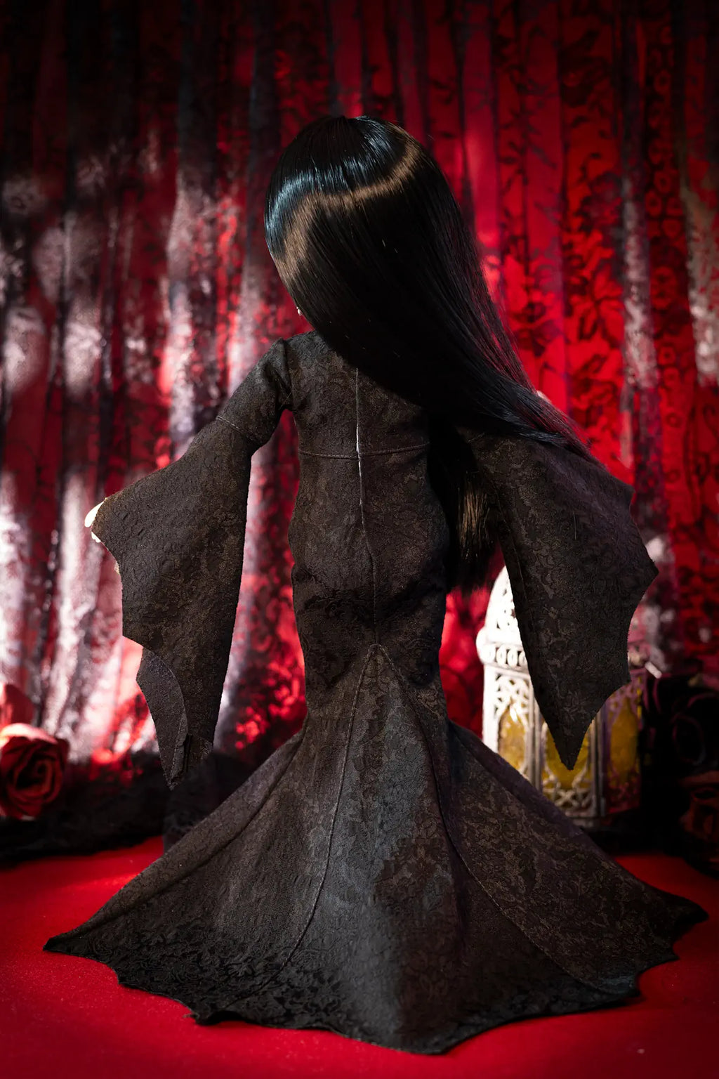 A doll or fashion figure shown from the back, wearing a long black mermaid-style gown with dramatic flared sleeves, posed against a dark red patterned curtain backdrop.