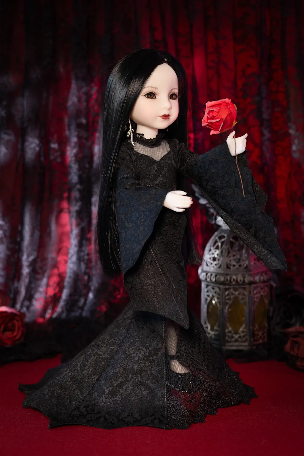 A gothic-style doll with long black hair and pale skin, wearing a black lace dress and holding a red rose, posed against a dark red patterned curtain backdrop.