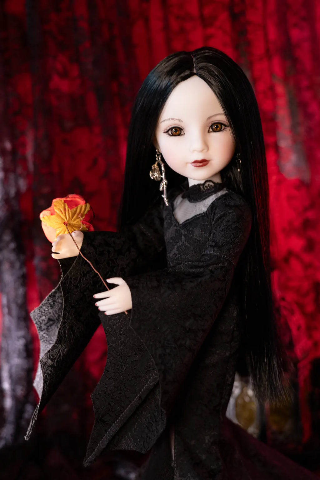 A doll with long black hair, pale skin, and dark eyes, wearing a black lace dress and holding a red and yellow flower, posed against a dark red patterned backdrop.