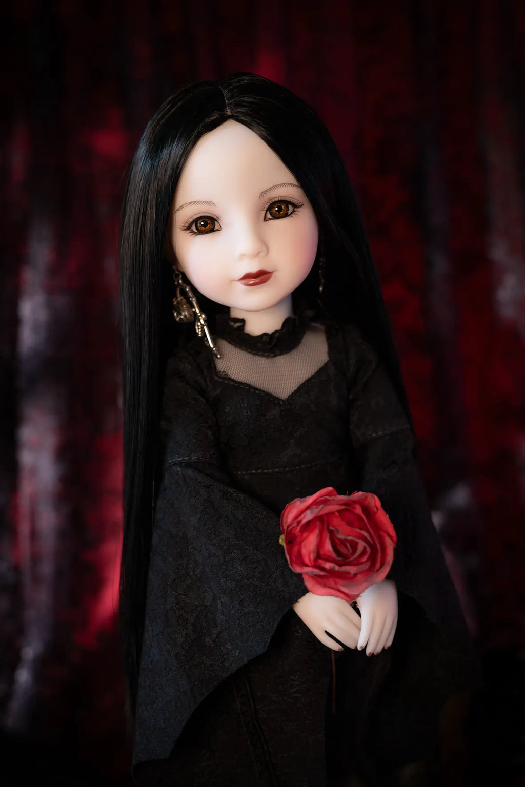 A gothic-style doll with long black hair, pale skin, and large brown eyes, wearing a black dress and holding a red rose against a dark red textured background.
