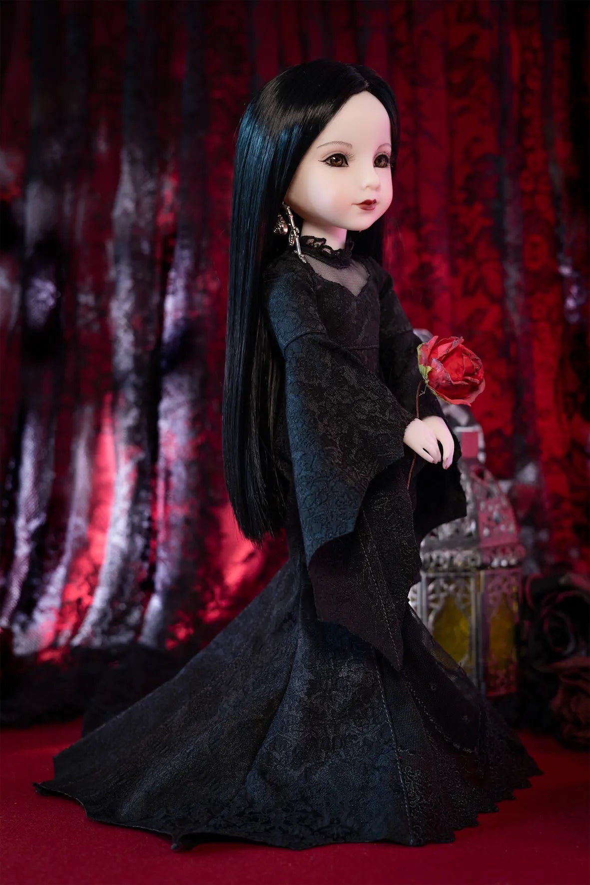 A doll with long black hair wearing a black lace dress, holding a red rose, posed against a dark red patterned backdrop.
