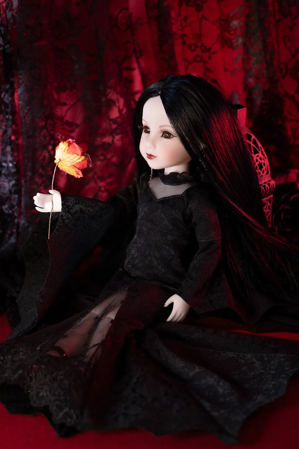 A doll with long black hair wearing a black dress, seated against a dark red patterned backdrop and holding a glowing orange flower.