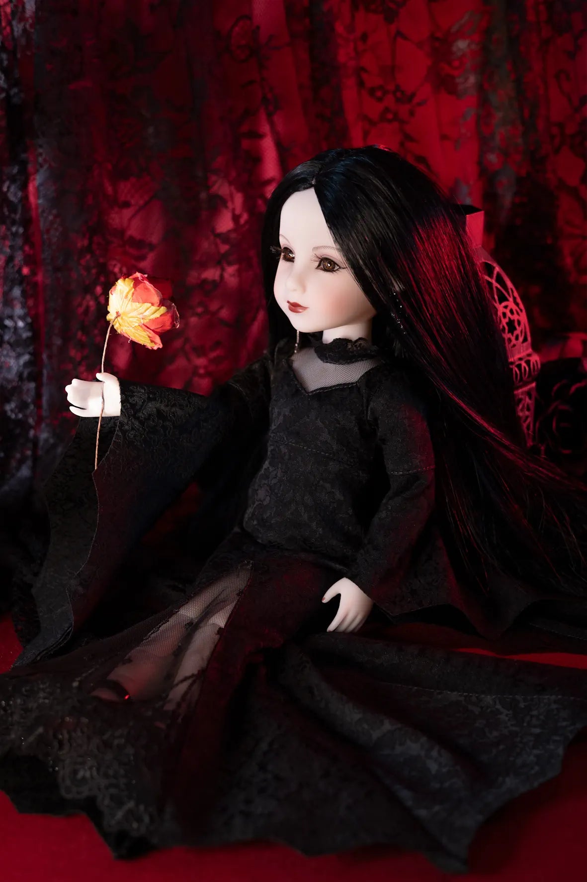 A doll with long black hair wearing a black dress, seated against a dark red patterned backdrop and holding a glowing orange flower.