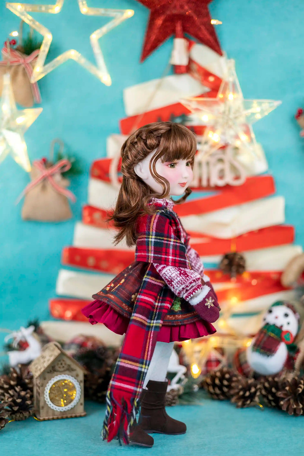 Doll in a plaid dress standing in front of a Christmas tree with lights and decorations.