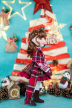Doll in a plaid dress standing in front of a Christmas tree with lights and decorations.