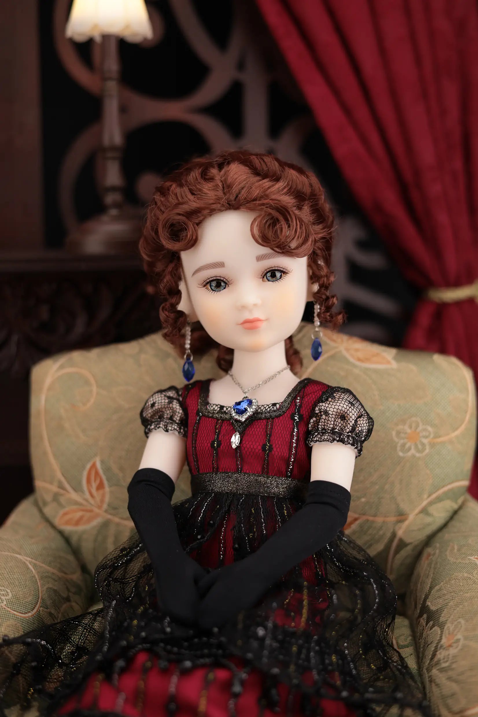 Doll in a red and black dress sitting on a decorative chair with a red curtain in the background.
