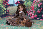Doll in a vintage outfit sitting on a patterned surface with flowers and a building in the background