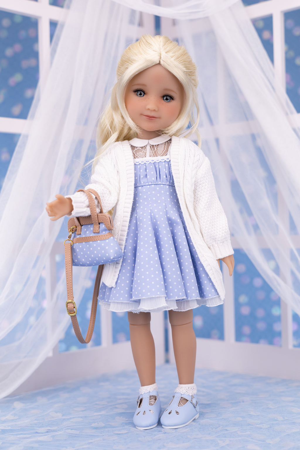 Doll wearing a blue dress with a white cardigan and holding a matching handbag against a decorative background.