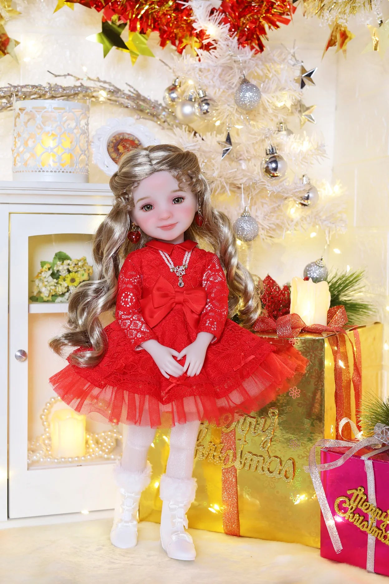Doll in a red dress standing in front of a decorated Christmas tree with lights and presents.