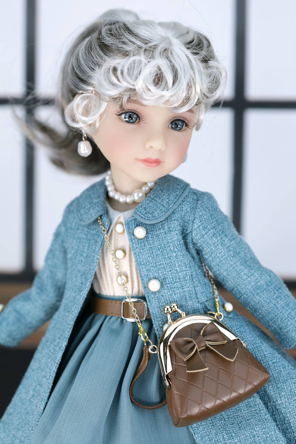 Doll wearing a dark turquoise coat with a brown handbag against a blurred background