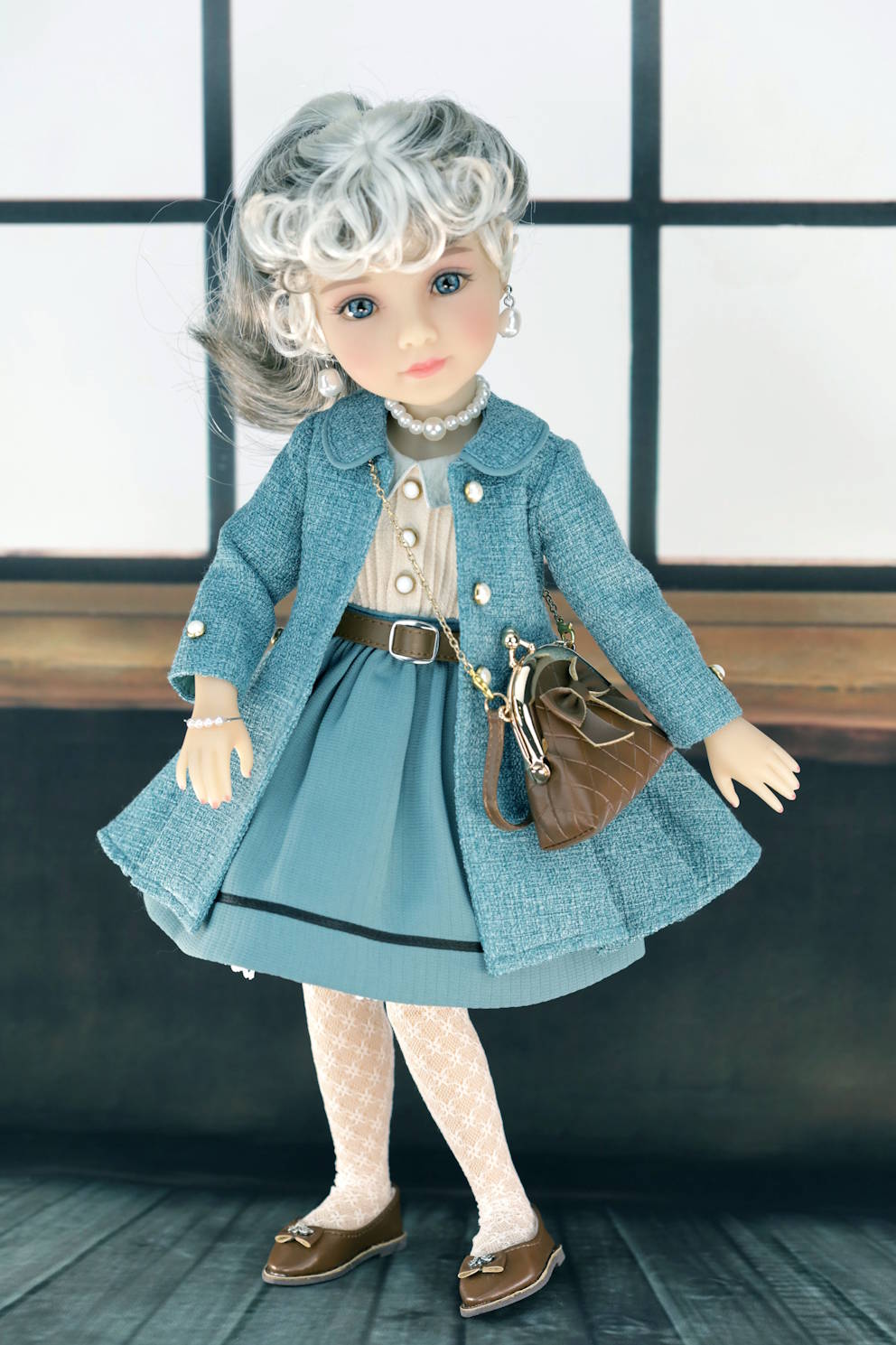 Doll wearing a dark turquoise coat and skirt with a brown bag against a neutral background