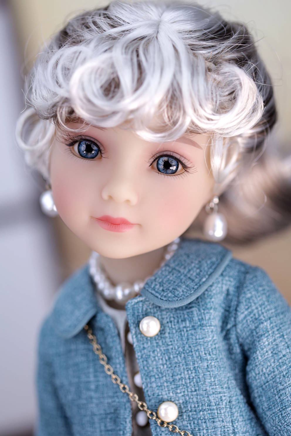 Close-up of a doll with white hair and blue eyes wearing a dark turquoise coat and pearl necklace.