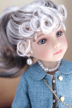 Close-up of a doll with white hair, blue eyes, and wearing a dark turquoise coat with a pearl necklace.