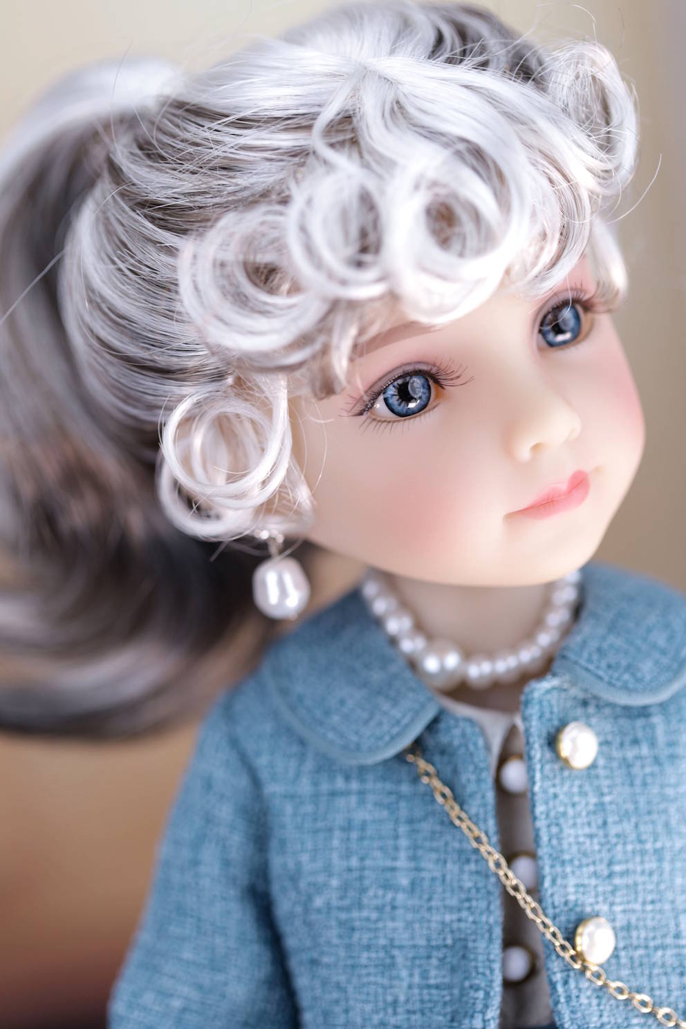 Close-up of a doll with white hair, blue eyes, and wearing a dark turquoise coat with a pearl necklace.