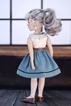 Doll with gray hair styled in an updo, wearing a light-colored top and blue skirt.