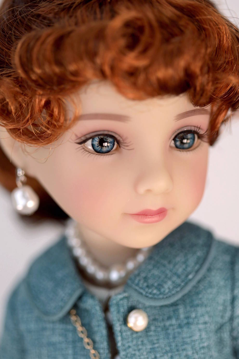 Close-up of a doll with red hair, blue eyes, wearing a dark turquoise jacket and pearl necklace.