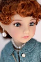 Close-up of a doll with red hair, blue eyes, wearing a dark turquoise jacket and pearl necklace.