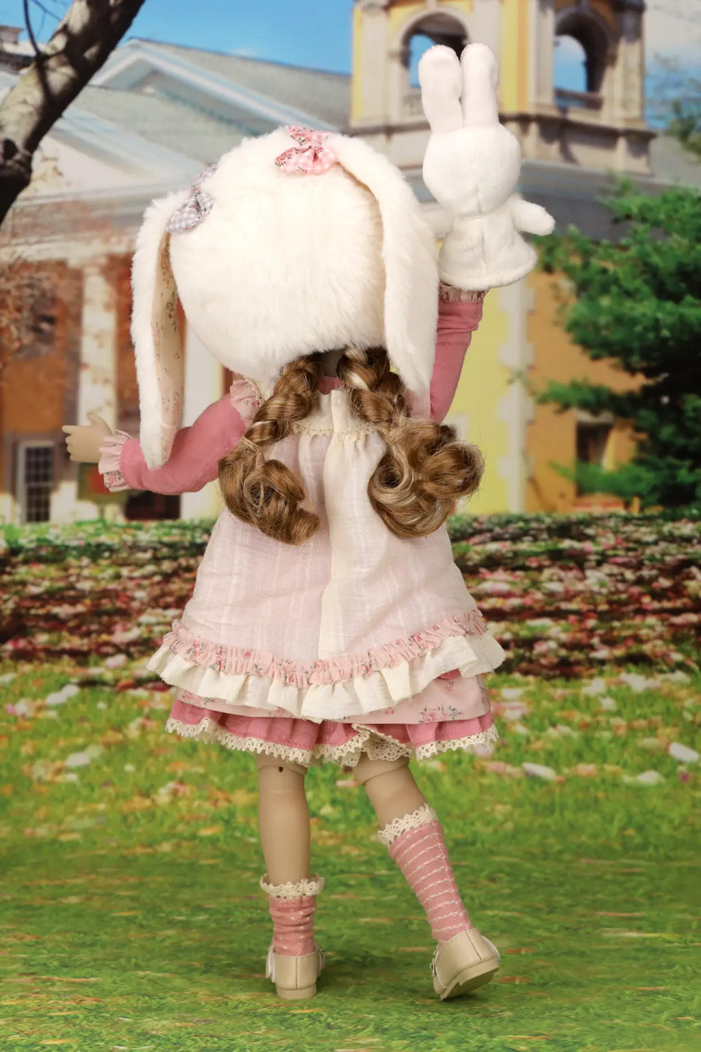 Doll in a pink dress with bunny ears holding large stuffed bunnies outdoors.