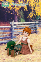 Doll in a brown dress with a green bag sitting on the ground with autumn leaves and a wooden fence in the background.