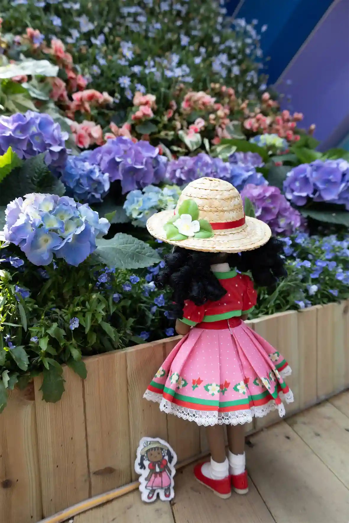Kyra in a pink dress and hat standing next to flowers and a wooden fence.
