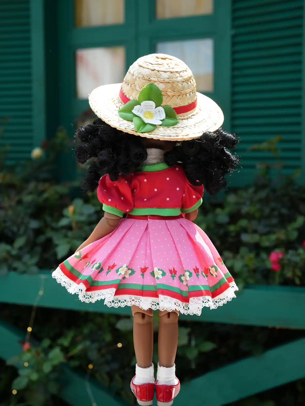 Kyra wearing a pink dress with red accents and a straw hat with floral decorations, standing in front of a green wooden door.