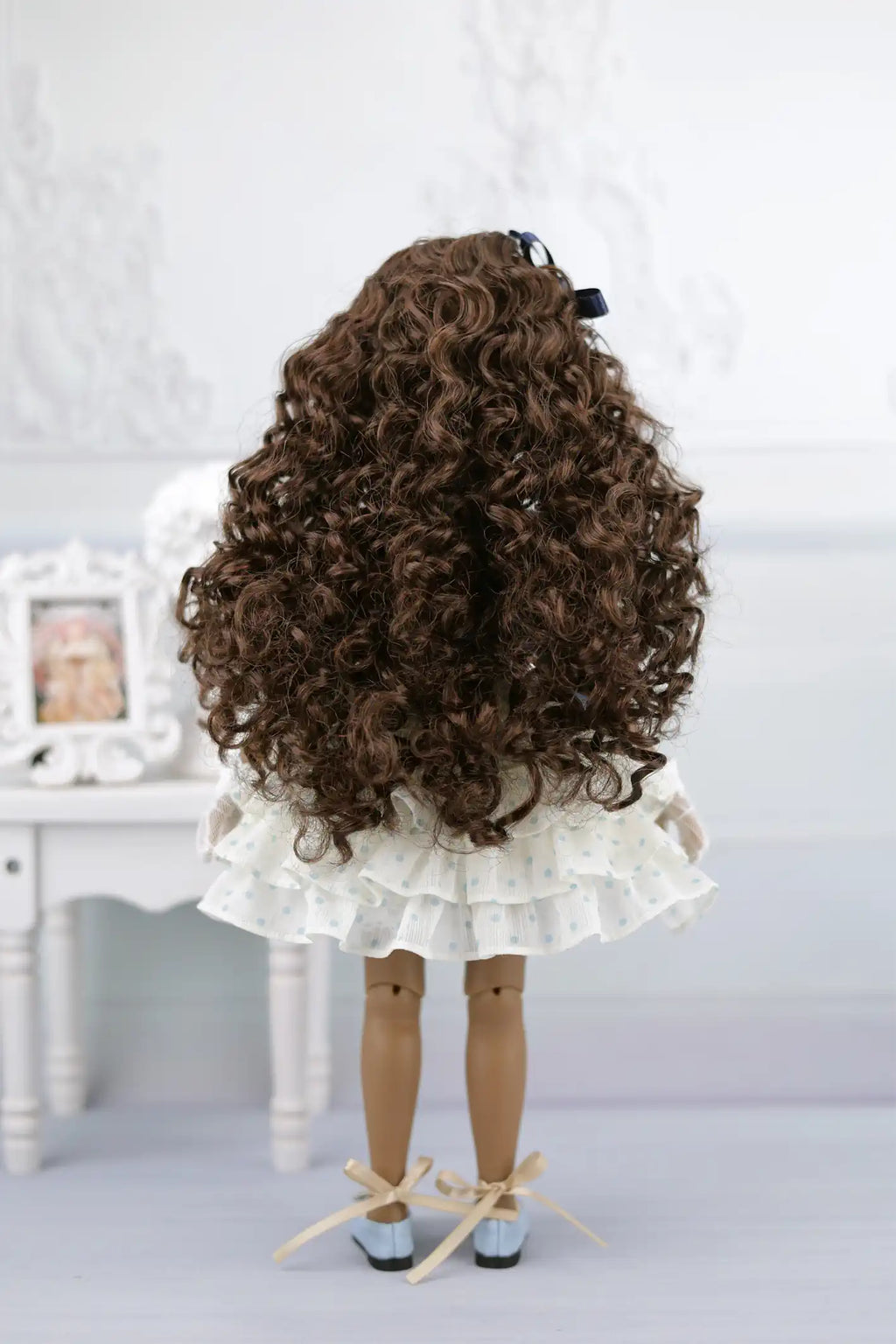 Ruth with curly hair and a white dress standing on a light gray surface.