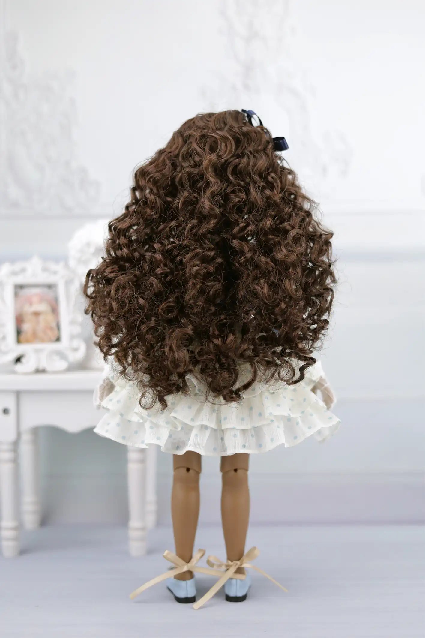 Ruth with curly hair and a white dress standing on a light gray surface.