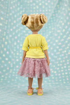 Juniper with blonde hair styled in buns, wearing a yellow top and pink lace skirt against a light blue background.