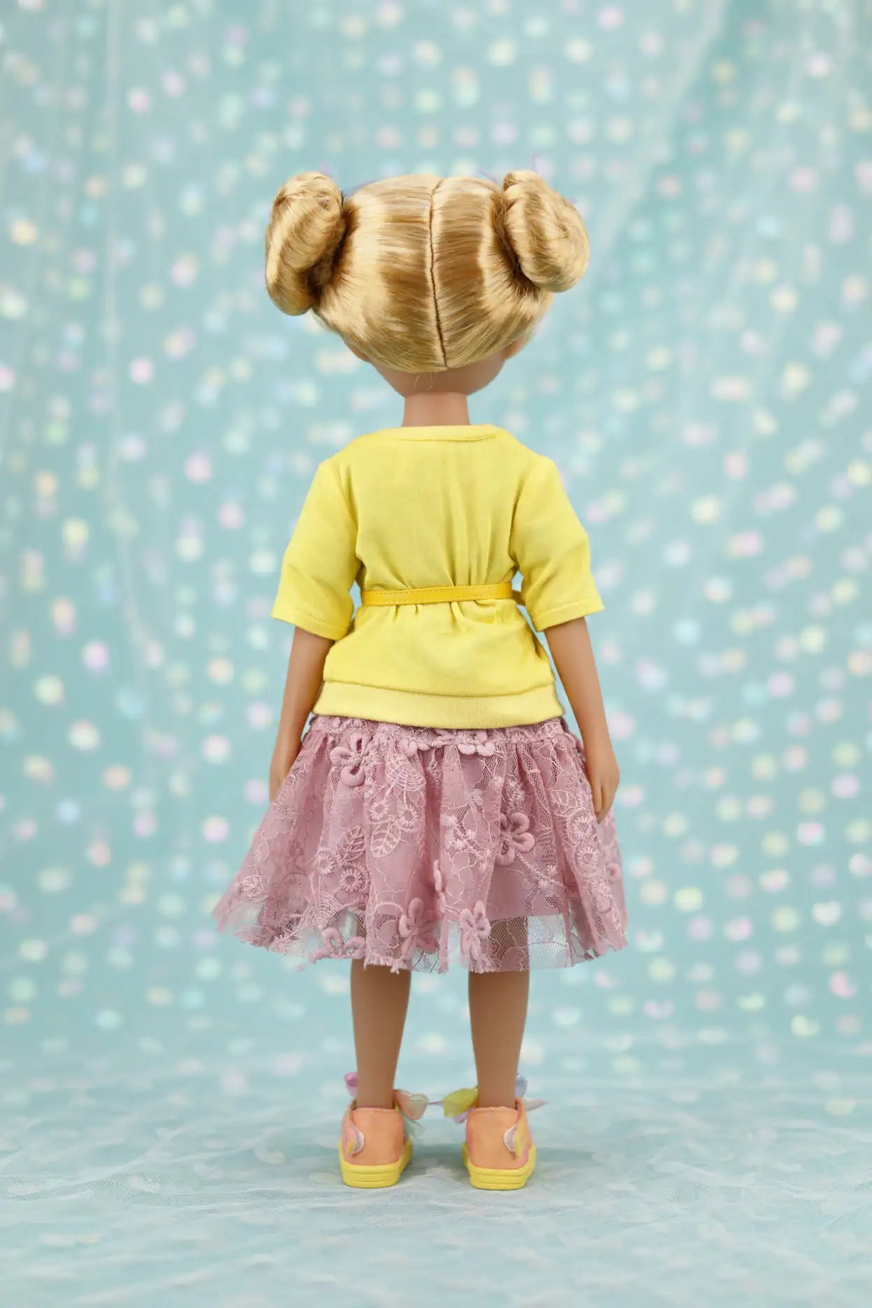 Juniper with blonde hair styled in buns, wearing a yellow top and pink lace skirt against a light blue background.