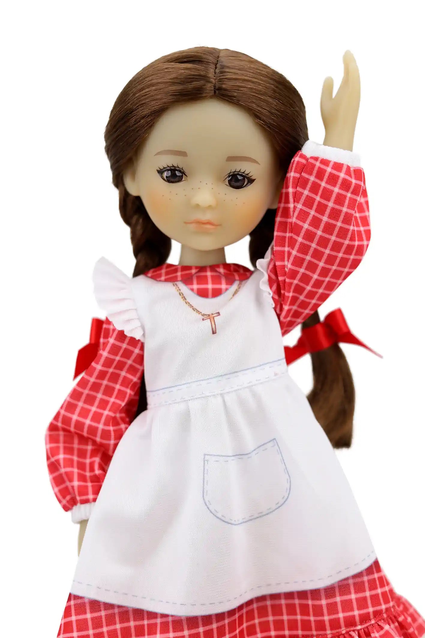 Jeannette, with brown braids and red bows, waves while dressed in a checkered outfit with a white apron and a cross pendant.