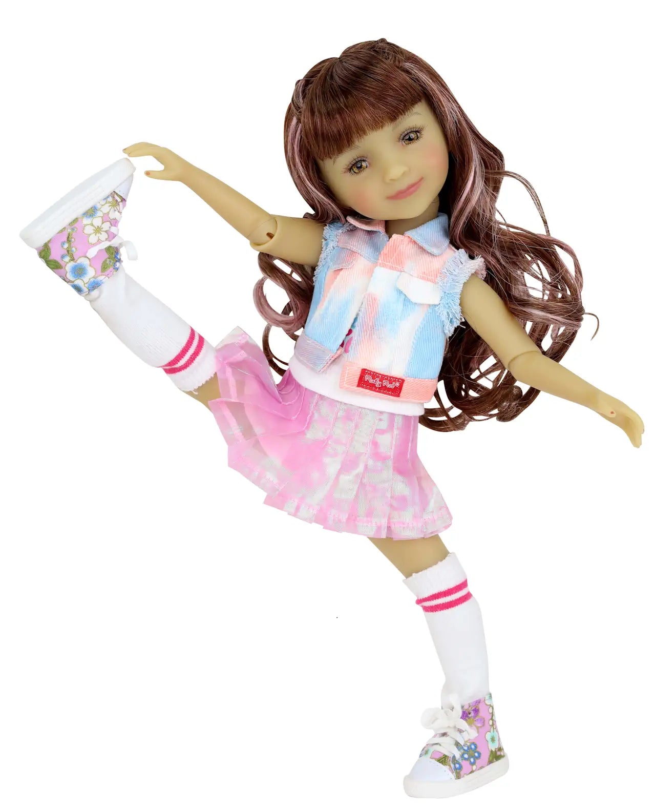 Bella in tie-dye vest, pink skirt, white socks with pink stripes, and floral sneakers, posing with one leg raised, white background.
