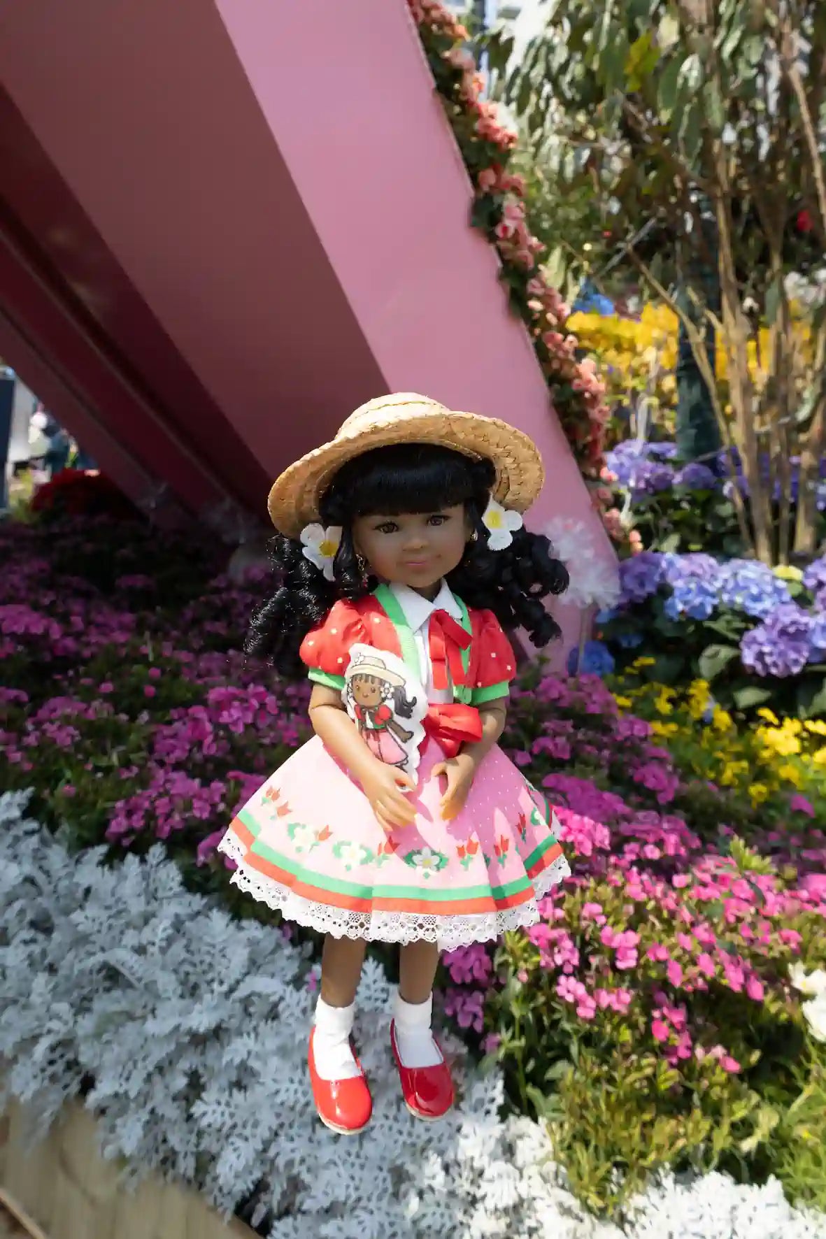 Kyra in a floral dress and hat standing among flowers and a pink structure.