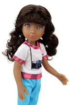 Mila, a doll with long curly brown hair, wears a white t-shirt with a camera graphic, light blue pants, and a camera strap, with one arm extended.