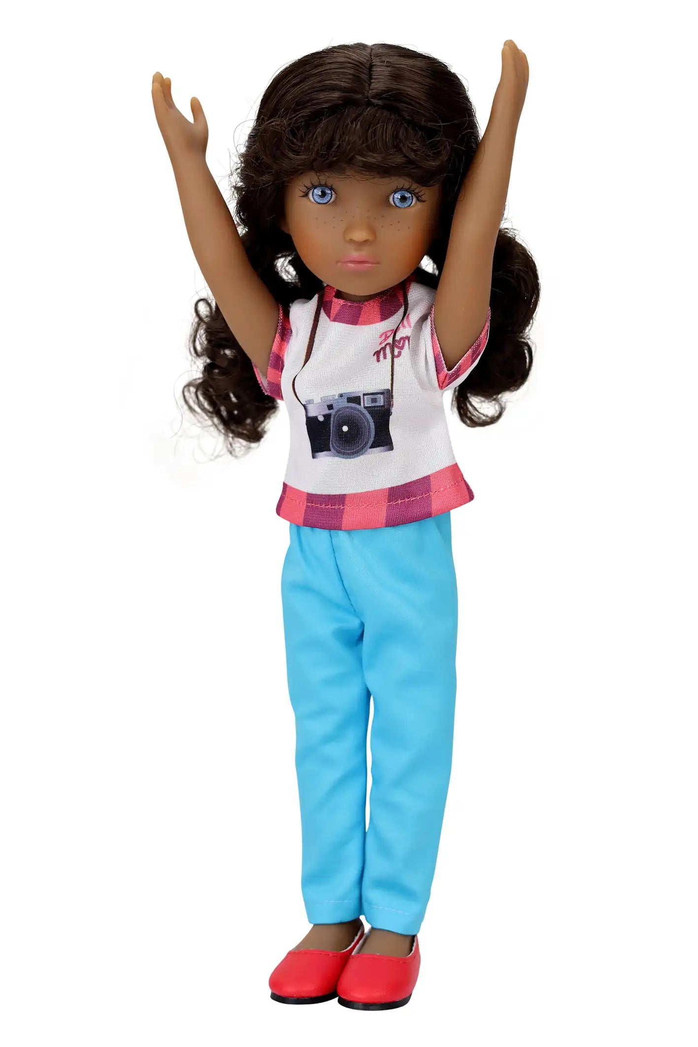 Mila, a doll with curly brown hair, wears a white shirt with a camera graphic, light blue pants, and red shoes, posing cheerfully with arms raised against a white background.