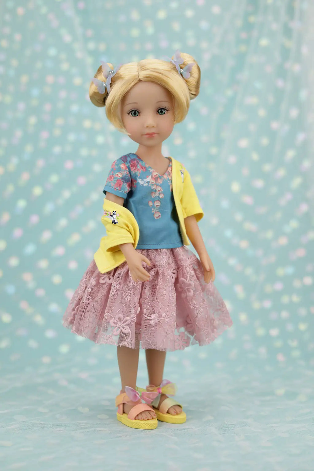 Juniper with blonde hair styled in buns, wearing a yellow cardigan over a blue shirt and pink skirt, on a light blue background with polka dots.