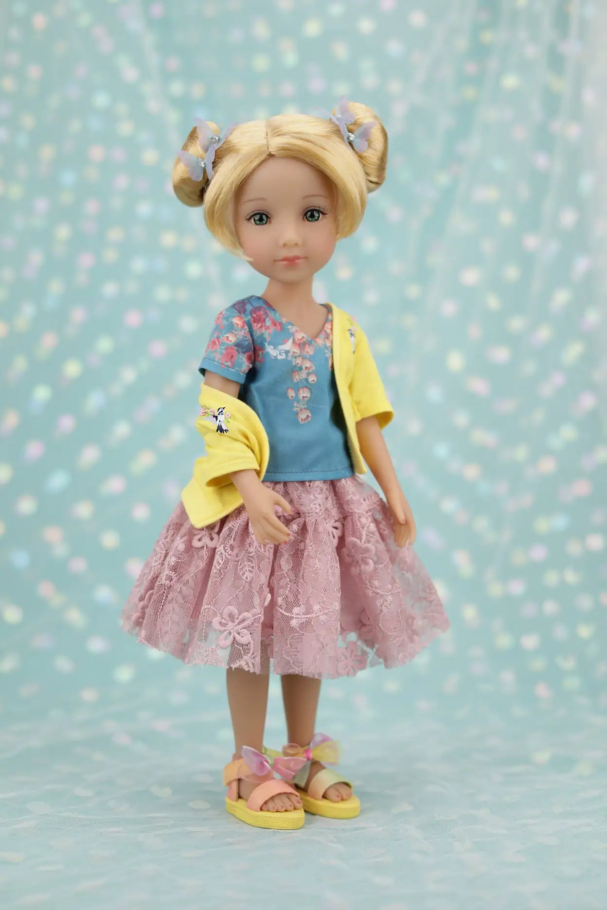 Juniper with blonde hair styled in buns, wearing a yellow cardigan over a blue shirt and pink skirt, on a light blue background with polka dots.