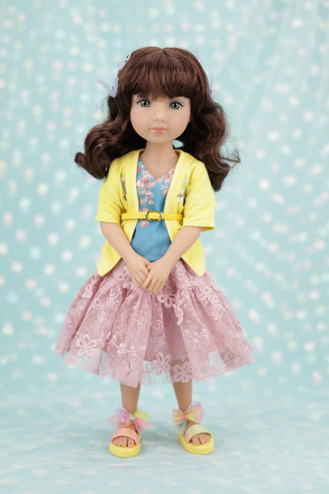 Juniper wearing a yellow cardigan, pink dress, and sandals on a light blue polka dot background