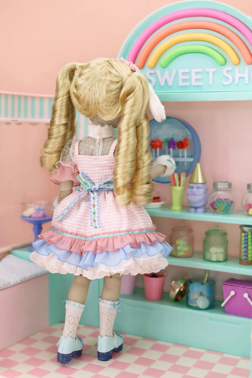 Doll in a pink dress standing in a colorful playroom with a rainbow and 'Sweet Shop' sign.