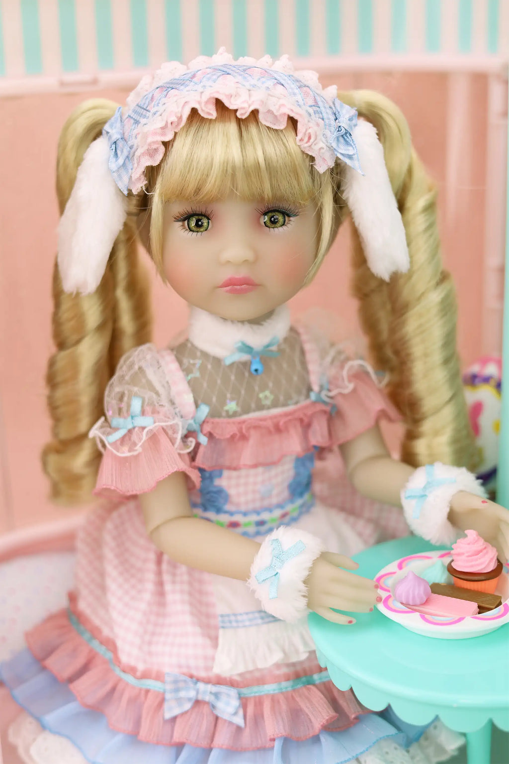 Doll with blonde hair and a detailed outfit sitting at a table with a pink background