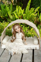 Doll in a white dress sitting under a canopy with greenery in the background