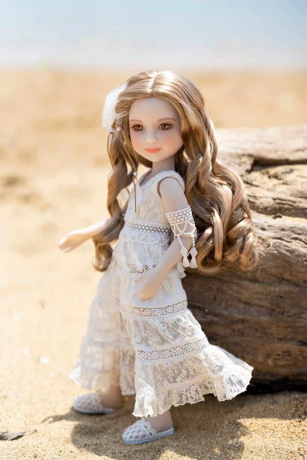Doll in a white dress sitting on a beach with sand and a log in the background