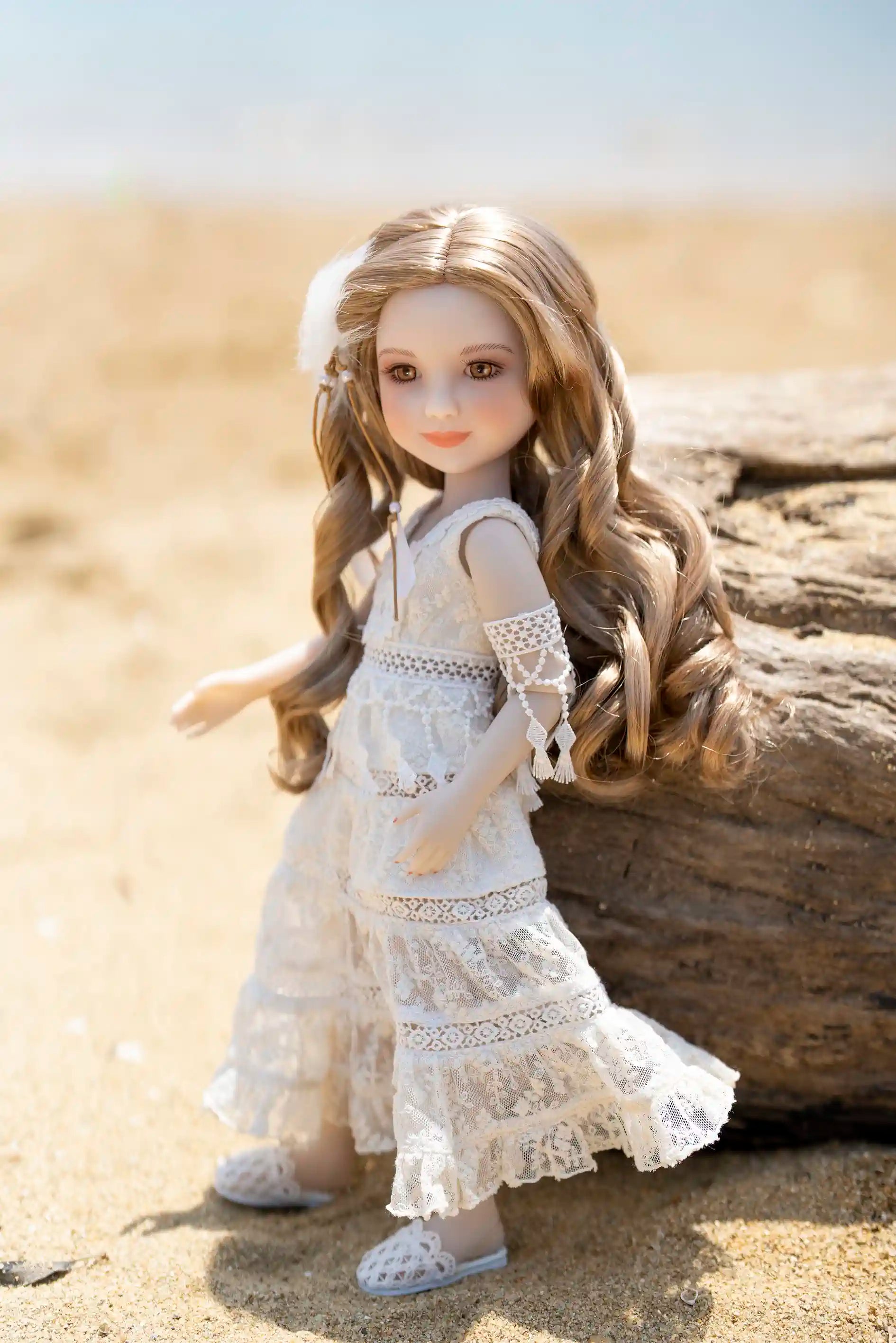 Doll in a white dress sitting on a beach with sand and a log in the background