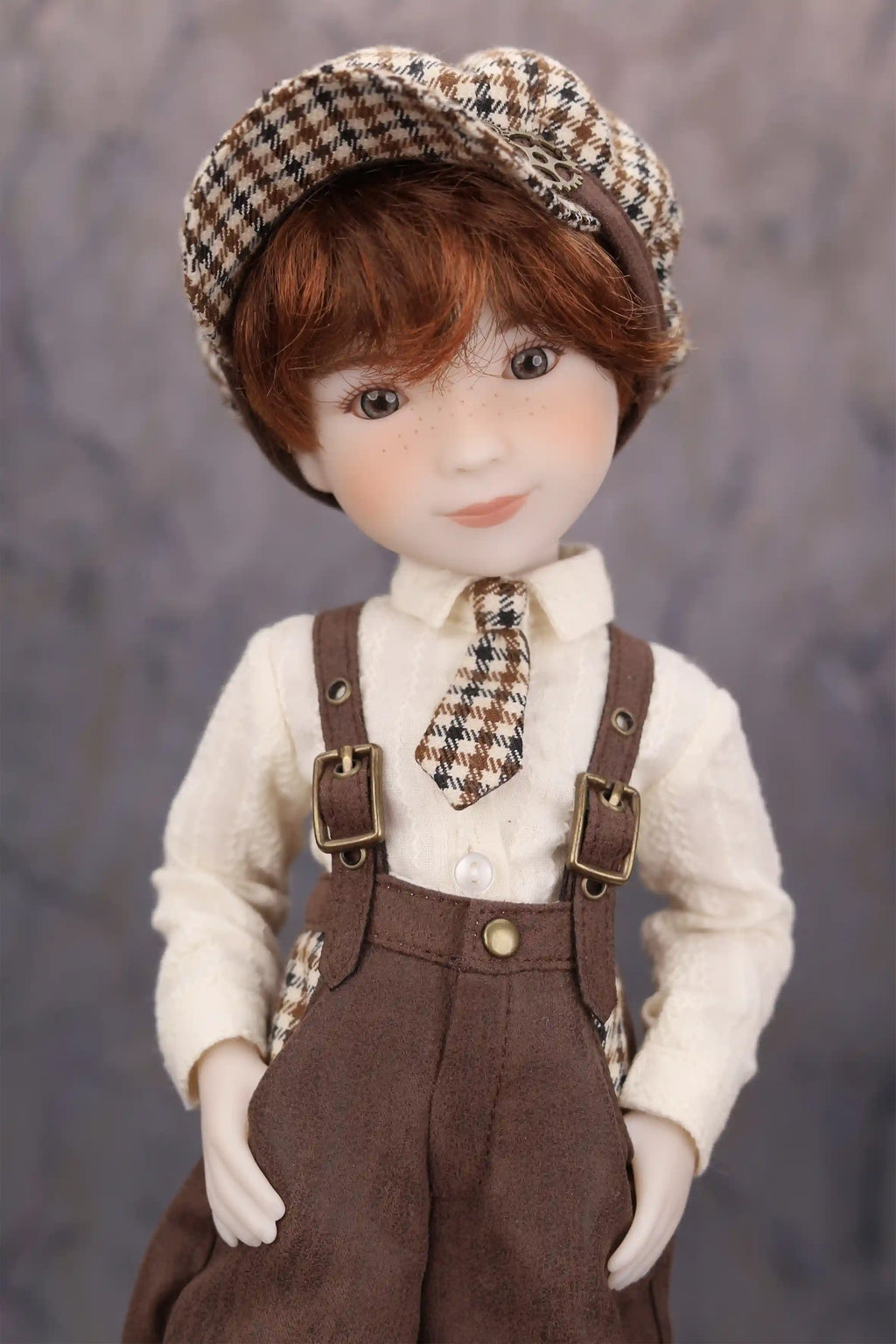 Doll wearing a checkered hat, white shirt and brown overalls against a gray background.