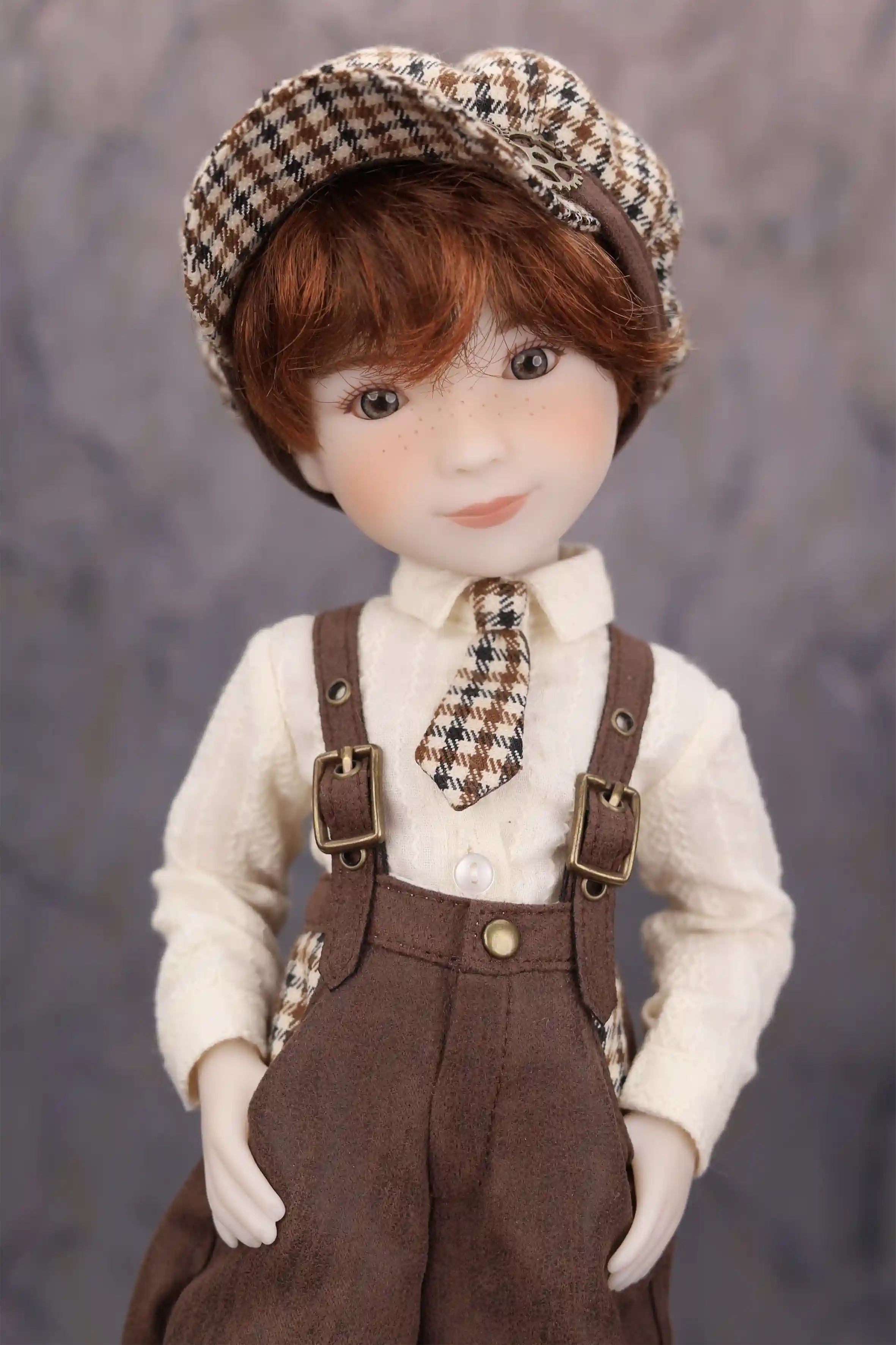 Doll wearing a checkered hat, white shirt and brown overalls against a gray background.