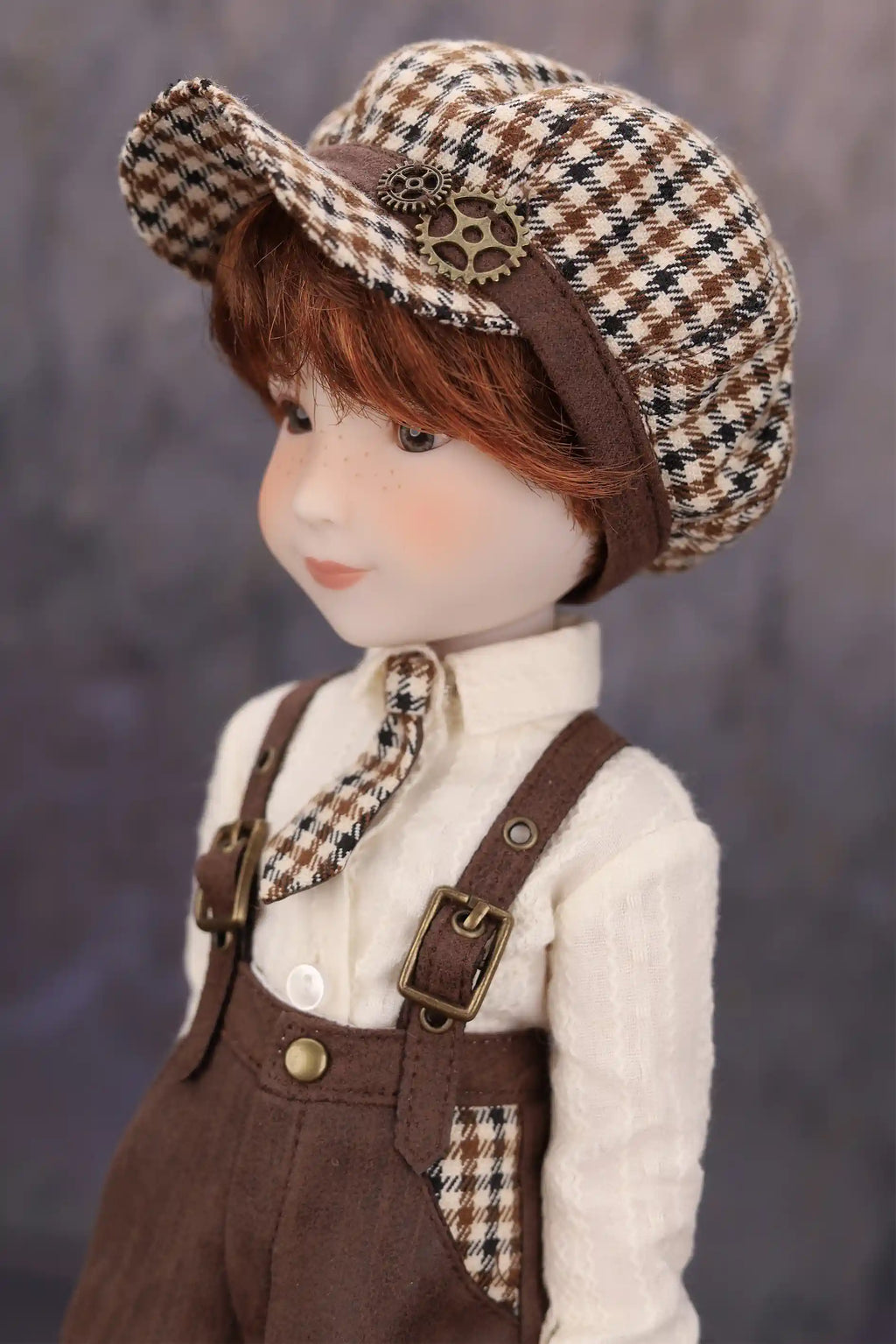 Doll wearing a checkered cap, white shirt, and brown overalls against a gary background