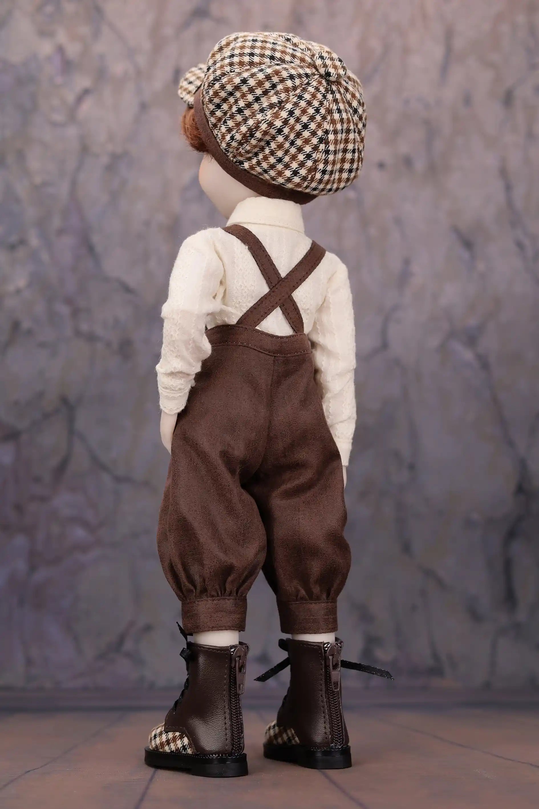 Doll wearing a checkered hat, white shirt, brown overalls, and black boots against a textured gray background.